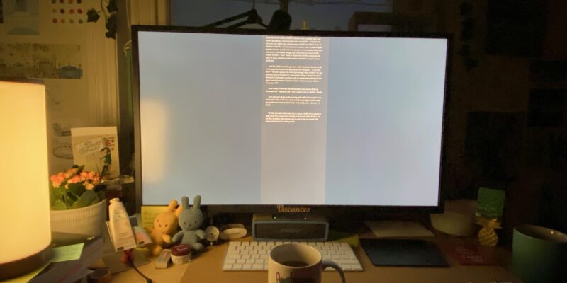 Desk with big monitor, lit from the left. A mug sits in front of the keyboard.