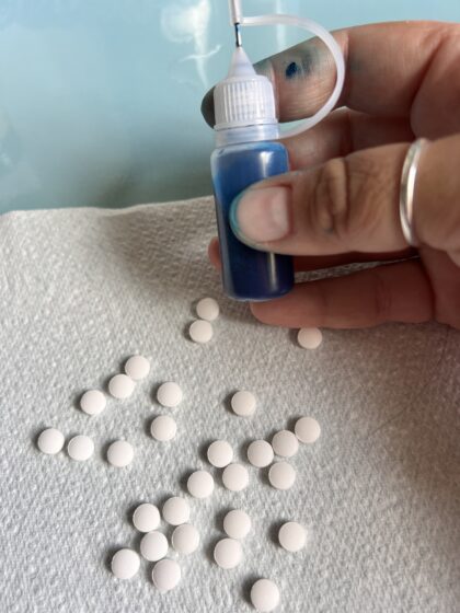 Tiny white pills on a kitchen towel, with a small bottle full of blue liquid being held above them.