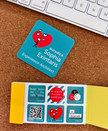 square business card "eurydice sophia exintaris experience architect" on desk next to keyboard and a sheet of stickers "be so good they can't ignore you", "build the right thing, build the thing right", "it depends", stick figure with floating hearts on a big red heart background