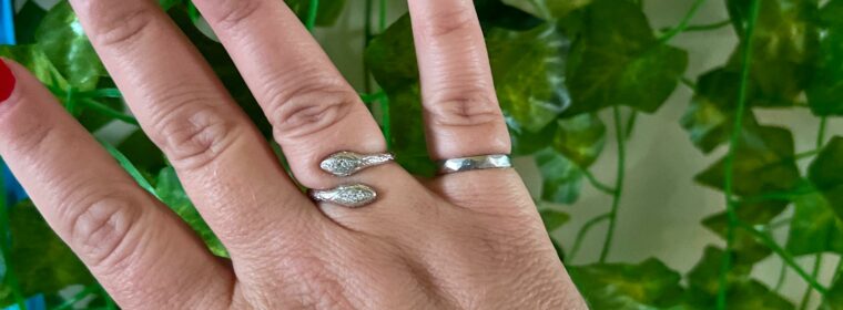 Right hand with iron right on the pinkie finger, painted red nails, a thumbring, and several blue and metal bracelets on the right wrist. The background is green leaves.
